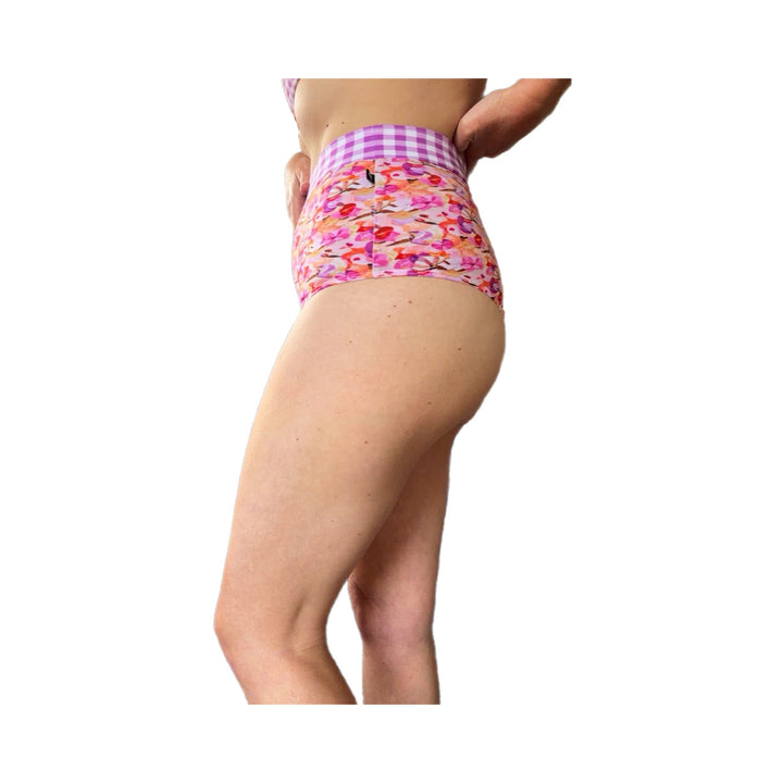 A side profile view of a model wearing high-waisted bikini bottoms with a pink and purple gingham waistband and a vibrant floral print body, set against a plain white background.