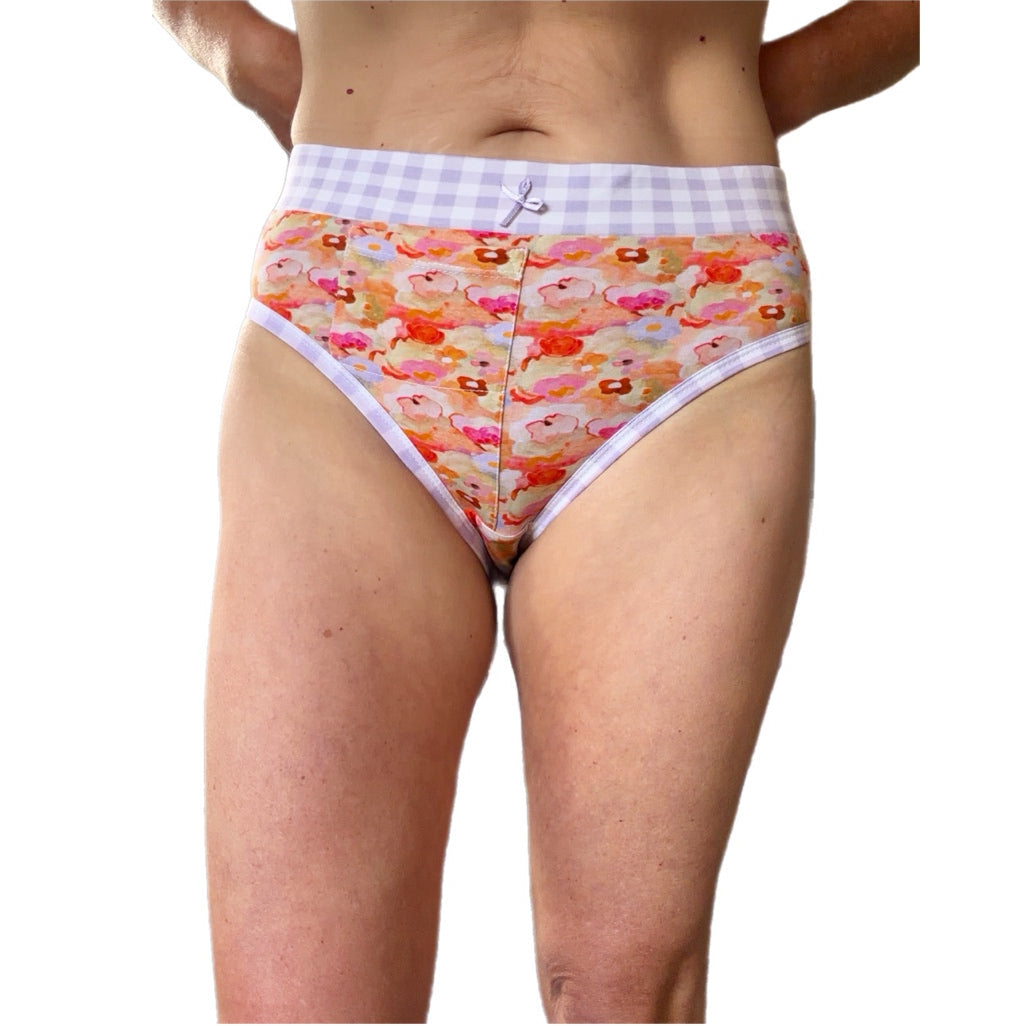 A woman wearing mid-rise briefs with a white and lavender gingham waistband and a colorful pastel floral print, shown from the waist to upper thighs on a plain white background.