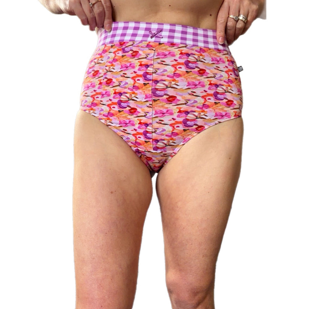 A model wearing high-waisted bikini bottoms with a colorful floral print and a purple-and-white gingham waistband, shown against a plain white background.